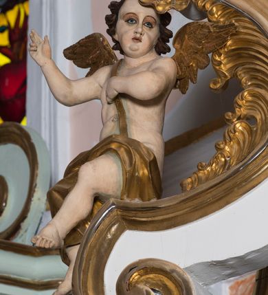 Zdjęcie nr 7: Para pełnoplastycznych, całopostaciowych figur aniołów o sylwetkach dziecięcych przysiadających na spływach wolutowych po obydwu stronach zwieńczenia ołtarza. Zwrócone są one niemal na wprost. Lekko przechylone w bok głowy charakteryzują się zaokrąglonymi twarzami o pełnych, wyrazistych, dziecięcych rysach. Krótkie, kręcone, ciemnobrązowe włosy ujmują twarze zebranymi po bokach i nad czołem puklami. Jedną rękę składają na piersiach, drugą unoszą bądź wyciągają w bok. Nagie sylwetki aniołów osłania w partii bioder złocona tkanina, a przez nagie torsy przewieszone są złocone szarfy. Na plecach mają złocone skrzydełka. 