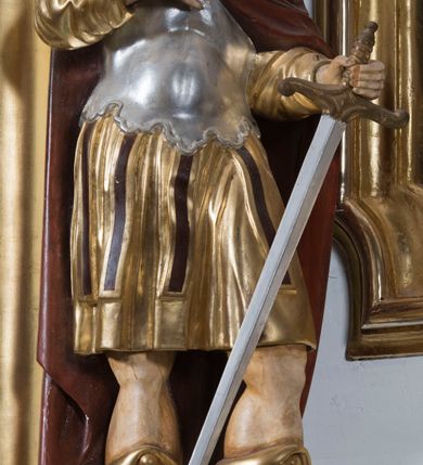 Zdjęcie nr 2: Całopostaciowa, drążona z tyłu figura przedstawia św. Kryspina stojącego w kontrapoście na czterobocznym, profilowanym postumencie z napisem „Ś(więty) Kryspin”. Święty zwrócony jest niemal profilem w prawo. Skierowaną w trzech czwartych w prawo, nieznacznie odchyloną do tyłu głowę charakteryzuje pociągła, owalna twarz z wyraziście i nieco sumarycznie opracowanymi rysami. Blada karnacja uwydatnia wyraziste oczy, długi, prosty wąski nos oraz niewielkie, pełne usta. Mocno skręcone długie, ciemnobrązowe włosy opadają na barki. Charakterystyczne są dwa, ujmujące twarz na wysokości uszu, pukle w formie ażurowych pętelek. Ubiór świętego, przypominający paradną zbroję żołnierza rzymskiego, składa się z krótkiej, sięgającej kolan, złoconej tuniki z długimi rękawami, osłaniającej tors, srebrzonej zbroi płytowej o dekoracyjnie wyciętej dolnej krawędzi oraz spiętego na prawym ramieniu purpurowego płaszcza. Na nogach są wysoko wiązane buty o wywiniętych cholewkach. Święty prawą rękę wspiera na piersiach, w lewej trzyma miecz o srebrzonej głowni.   