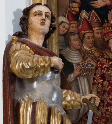 Zdjęcie nr 1: Całopostaciowa, drążona z tyłu figura przedstawia św. Kryspina stojącego w kontrapoście na czterobocznym, profilowanym postumencie z napisem „Ś(więty) Kryspin”. Święty zwrócony jest niemal profilem w prawo. Skierowaną w trzech czwartych w prawo, nieznacznie odchyloną do tyłu głowę charakteryzuje pociągła, owalna twarz z wyraziście i nieco sumarycznie opracowanymi rysami. Blada karnacja uwydatnia wyraziste oczy, długi, prosty wąski nos oraz niewielkie, pełne usta. Mocno skręcone długie, ciemnobrązowe włosy opadają na barki. Charakterystyczne są dwa, ujmujące twarz na wysokości uszu, pukle w formie ażurowych pętelek. Ubiór świętego, przypominający paradną zbroję żołnierza rzymskiego, składa się z krótkiej, sięgającej kolan, złoconej tuniki z długimi rękawami, osłaniającej tors, srebrzonej zbroi płytowej o dekoracyjnie wyciętej dolnej krawędzi oraz spiętego na prawym ramieniu purpurowego płaszcza. Na nogach są wysoko wiązane buty o wywiniętych cholewkach. Święty prawą rękę wspiera na piersiach, w lewej trzyma miecz o srebrzonej głowni.   