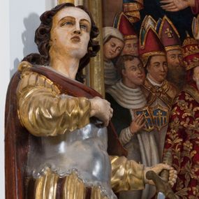 Zdjęcie nr 1: Całopostaciowa, drążona z tyłu figura przedstawia św. Kryspina stojącego w kontrapoście na czterobocznym, profilowanym postumencie z napisem „Ś(więty) Kryspin”. Święty zwrócony jest niemal profilem w prawo. Skierowaną w trzech czwartych w prawo, nieznacznie odchyloną do tyłu głowę charakteryzuje pociągła, owalna twarz z wyraziście i nieco sumarycznie opracowanymi rysami. Blada karnacja uwydatnia wyraziste oczy, długi, prosty wąski nos oraz niewielkie, pełne usta. Mocno skręcone długie, ciemnobrązowe włosy opadają na barki. Charakterystyczne są dwa, ujmujące twarz na wysokości uszu, pukle w formie ażurowych pętelek. Ubiór świętego, przypominający paradną zbroję żołnierza rzymskiego, składa się z krótkiej, sięgającej kolan, złoconej tuniki z długimi rękawami, osłaniającej tors, srebrzonej zbroi płytowej o dekoracyjnie wyciętej dolnej krawędzi oraz spiętego na prawym ramieniu purpurowego płaszcza. Na nogach są wysoko wiązane buty o wywiniętych cholewkach. Święty prawą rękę wspiera na piersiach, w lewej trzyma miecz o srebrzonej głowni.   