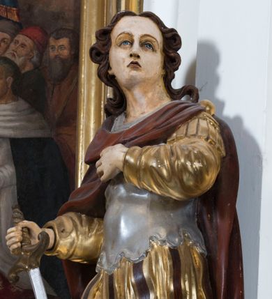 Zdjęcie nr 1: Całopostaciowa, drążona z tyłu figura przedstawia św. Kryspiniana stojącego w kontrapoście na czterobocznym, profilowanym postumencie z napisem „Ś(więty) Kryspinian”. Święty zwrócony jest niemal profilem w lewo. Skierowaną na wprost, uniesioną głowę charakteryzuje owalna twarz z wyraziście i nieco sumarycznie opracowanymi, młodzieńczymi rysami. Blada karnacja uwydatnia ciemną oprawę oczu, długi, prosty wąski nos oraz niewielkie, pełne usta. Mocno skręcone długie, ciemnobrązowe włosy dekoracyjnie ufryzowanymi puklami opadają na barki. Ubiór świętego, przypominający paradną zbroję żołnierza rzymskiego, składa się z krótkiej, sięgającej kolan, złoconej tuniki z długimi rękawami, osłaniającej tors, srebrzonej zbroi płytowej o dekoracyjnie wyciętej dolnej krawędzi oraz spiętego na lewym ramieniu purpurowego płaszcza. Na nogach są wysoko wiązane buty o wywiniętych cholewkach. Święty lewą rękę wspiera na piersiach, w prawej trzyma miecz o srebrzonej głowni.   