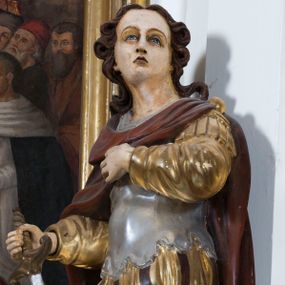 Zdjęcie nr 1: Całopostaciowa, drążona z tyłu figura przedstawia św. Kryspiniana stojącego w kontrapoście na czterobocznym, profilowanym postumencie z napisem „Ś(więty) Kryspinian”. Święty zwrócony jest niemal profilem w lewo. Skierowaną na wprost, uniesioną głowę charakteryzuje owalna twarz z wyraziście i nieco sumarycznie opracowanymi, młodzieńczymi rysami. Blada karnacja uwydatnia ciemną oprawę oczu, długi, prosty wąski nos oraz niewielkie, pełne usta. Mocno skręcone długie, ciemnobrązowe włosy dekoracyjnie ufryzowanymi puklami opadają na barki. Ubiór świętego, przypominający paradną zbroję żołnierza rzymskiego, składa się z krótkiej, sięgającej kolan, złoconej tuniki z długimi rękawami, osłaniającej tors, srebrzonej zbroi płytowej o dekoracyjnie wyciętej dolnej krawędzi oraz spiętego na lewym ramieniu purpurowego płaszcza. Na nogach są wysoko wiązane buty o wywiniętych cholewkach. Święty lewą rękę wspiera na piersiach, w prawej trzyma miecz o srebrzonej głowni.   