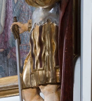 Zdjęcie nr 2: Całopostaciowa, drążona z tyłu figura przedstawia św. Kryspiniana stojącego w kontrapoście na czterobocznym, profilowanym postumencie z napisem „Ś(więty) Kryspinian”. Święty zwrócony jest niemal profilem w lewo. Skierowaną na wprost, uniesioną głowę charakteryzuje owalna twarz z wyraziście i nieco sumarycznie opracowanymi, młodzieńczymi rysami. Blada karnacja uwydatnia ciemną oprawę oczu, długi, prosty wąski nos oraz niewielkie, pełne usta. Mocno skręcone długie, ciemnobrązowe włosy dekoracyjnie ufryzowanymi puklami opadają na barki. Ubiór świętego, przypominający paradną zbroję żołnierza rzymskiego, składa się z krótkiej, sięgającej kolan, złoconej tuniki z długimi rękawami, osłaniającej tors, srebrzonej zbroi płytowej o dekoracyjnie wyciętej dolnej krawędzi oraz spiętego na lewym ramieniu purpurowego płaszcza. Na nogach są wysoko wiązane buty o wywiniętych cholewkach. Święty lewą rękę wspiera na piersiach, w prawej trzyma miecz o srebrzonej głowni.   