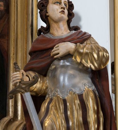 Zdjęcie nr 4: Całopostaciowa, drążona z tyłu figura przedstawia św. Kryspiniana stojącego w kontrapoście na czterobocznym, profilowanym postumencie z napisem „Ś(więty) Kryspinian”. Święty zwrócony jest niemal profilem w lewo. Skierowaną na wprost, uniesioną głowę charakteryzuje owalna twarz z wyraziście i nieco sumarycznie opracowanymi, młodzieńczymi rysami. Blada karnacja uwydatnia ciemną oprawę oczu, długi, prosty wąski nos oraz niewielkie, pełne usta. Mocno skręcone długie, ciemnobrązowe włosy dekoracyjnie ufryzowanymi puklami opadają na barki. Ubiór świętego, przypominający paradną zbroję żołnierza rzymskiego, składa się z krótkiej, sięgającej kolan, złoconej tuniki z długimi rękawami, osłaniającej tors, srebrzonej zbroi płytowej o dekoracyjnie wyciętej dolnej krawędzi oraz spiętego na lewym ramieniu purpurowego płaszcza. Na nogach są wysoko wiązane buty o wywiniętych cholewkach. Święty lewą rękę wspiera na piersiach, w prawej trzyma miecz o srebrzonej głowni.   