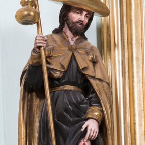 Zdjęcie nr 1: Całopostaciowa, pełnoplastyczna rzeźba przedstawia stojącego na niewysokiej, prostopadłościennej podstawie o ściętych narożach św. Rocha zwróconego na wprost. Głowa i ramiona mężczyzny skierowane są w trzech czwartych w prawo. Owalną twarz charakteryzują łagodne rysy dojrzałego mężczyzny z szeroko rozstawionymi, lekko przymkniętymi oczami z łagodnymi łukami wyrazistych brwi, długim, spiczastym nosem oraz krótkim zarostem. Na barki opadają długie, lekko falowane, ciemnobrązowe włosy. Święty ubrany jest w długą, ciemnobrązową, spiętą złoconym paskiem suknię ze złoconymi, podwiniętymi mankietami oraz spięty pod szyją kwadratową zaponą, długi, złocony płaszcz. Na głowę mężczyzny nałożony jest złocony kapelusz z lekko wywiniętym z przodu, szerokim rondem. Na nogach ma złocone buty z wysokimi cholewkami. W prawej dłoni trzyma kij pielgrzymi o złoconym drzewcu z zawieszoną na nim takąż tykwą. Lewą ręką unosi suknię wskazując jednocześnie na czerwoną ranę na udzie lewej, wspartej na niewielkim kamieniu nogi. 