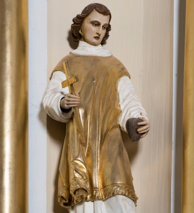 Zdjęcie nr 1: Całopostaciowa, drążona z tyłu rzeźba przedstawia stojącą w kontrapoście, na profilowanym postumencie z napisem „Ś(więty) Szczepan”, postać św. Szczepana męczennika zwróconego na wprost. Skierowaną w trzech czwartych w prawo, lekko pochyloną głowę charakteryzuje owalna twarz z sumarycznie opracowanymi, wyrazistymi, młodzieńczymi rysami o nieco przymkniętych oczach, długim nosie i drobnych ustach. Brązowe, sięgające szyi i podkręcone na końcach włosy opadają półkoliście ufryzowaną grzywką na czoło. Święty ma na sobie albę i złoconą dalmatykę, a na stopach jasnobrązowe buty. W prawej dłoni trzyma złocony krzyż, w lewej kamień.  