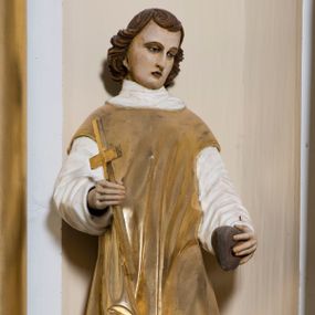 Zdjęcie nr 1: Całopostaciowa, drążona z tyłu rzeźba przedstawia stojącą w kontrapoście, na profilowanym postumencie z napisem „Ś(więty) Szczepan”, postać św. Szczepana męczennika zwróconego na wprost. Skierowaną w trzech czwartych w prawo, lekko pochyloną głowę charakteryzuje owalna twarz z sumarycznie opracowanymi, wyrazistymi, młodzieńczymi rysami o nieco przymkniętych oczach, długim nosie i drobnych ustach. Brązowe, sięgające szyi i podkręcone na końcach włosy opadają półkoliście ufryzowaną grzywką na czoło. Święty ma na sobie albę i złoconą dalmatykę, a na stopach jasnobrązowe buty. W prawej dłoni trzyma złocony krzyż, w lewej kamień.  