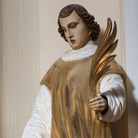 Zdjęcie nr 1: Całopostaciowa, drążona z tyłu rzeźba przedstawia stojącego w kontrapoście św. Wawrzyńca zwróconego na wprost, dodatkowo sylwetkę charakteryzuje delikatny układ „figura serpentinata”. Pochyloną głowę cechuje owalna w obrysie twarz o sumarycznie opracowanych, wyrazistych, młodzieńczych rysach, z długim, prostym nosem i przymkniętymi oczami. Skręcona w przylegający do głowy pukiel grzywka opada półkoliście na czoło, po bokach twarz ujmują podkręcone na końcach pasma sięgających szyi, ciemnobrązowych włosów. Święty ubrany jest w albę i złoconą dalmatykę, na stopach ma buty. Ręce wysunięte są nieznacznie do przodu. W uniesionej nieco do góry lewej dłoni trzyma złoconą gałązkę palmową, w prawej bliżej nieokreślony przedmiot (być może jest to uchwyt stojącego obok, złoconego rusztu).  