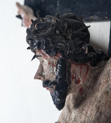 Zdjęcie nr 6: Do prostego krzyża przybita trzema gwoźdźmi pełnoplastyczna figura Chrystusa umarłego. Ciało w lekkim zwisie, o szeroko rozłożonych ramionach, głowa opadająca na prawy bark, nogi delikatnie ugięte w kolanach, stopy w układzie prawa na lewą. Chrystus ma podłużną twarz, z zamkniętymi oczami, długim, spiczastym nosem i wąskimi ustami, okoloną krótką, kręconą brodą. Włosy długie, ciemnobrązowe, silnie skręcone w pukle, opadające na plecy i prawe ramię. Na głowie szeroka korona cierniowa. Ciało szczupłe, o wyraźnie podkreślonej talii i szerokich biodrach, z delikatnie zaznaczoną muskulaturą i żebrami. Perizonium krótkie, odsłaniające prawe udo, zawiązane na prawym biodrze z opadającym wzdłuż nogi zwisem tkaniny. Polichromia ciała naturalistyczna, z zaznaczonymi śladami męki. Na zakończeniu pionowej belki krzyża kartusz z napisem „INRI”.
 
