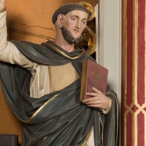 Zdjęcie nr 1: Pełnoplastyczna figura św. Dominika została ustawiona na niskim prostopadłościennym postumencie o ściętych narożach. Święty jest przedstawiony frontalnie w całej postaci w lekkim kontrapoście. Prawą rękę unosi w geście mowy, w lewej zaś na wysokości piersi trzyma księgę Ewangelii w ciemnoczerwonej oprawie zdobionej złotym krzyżem. Święty Dominik ma pociągłą twarz okoloną brązowym, krótkim zarostem oraz krótkimi włosami z widoczną tonsurą. Twarz o naturalistycznej karnacji jest drobna, z prostym nosem, wąskimi ustami oraz niebieskimi oczami. Zakonnik ubrany jest w biały dominikański habit opadający ostro drapowanymi fałdami ku ziemi, odsłaniający czubki szarych butów, jasnoochrowy szkaplerz lamowany złotym paskiem oraz szaroczarny płaszcz z kapturem narzucony na ramiona, lamowany złotym paskiem. Jego prawą połę święty podtrzymuje lewą ręką.  