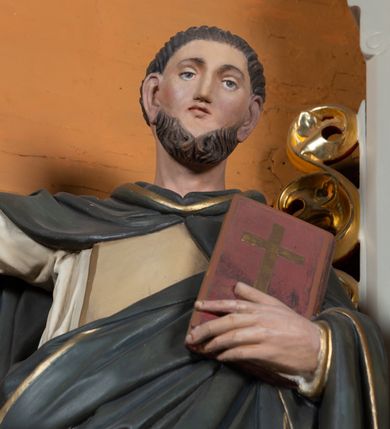 Zdjęcie nr 4: Pełnoplastyczna figura św. Dominika została ustawiona na niskim prostopadłościennym postumencie o ściętych narożach. Święty jest przedstawiony frontalnie w całej postaci w lekkim kontrapoście. Prawą rękę unosi w geście mowy, w lewej zaś na wysokości piersi trzyma księgę Ewangelii w ciemnoczerwonej oprawie zdobionej złotym krzyżem. Święty Dominik ma pociągłą twarz okoloną brązowym, krótkim zarostem oraz krótkimi włosami z widoczną tonsurą. Twarz o naturalistycznej karnacji jest drobna, z prostym nosem, wąskimi ustami oraz niebieskimi oczami. Zakonnik ubrany jest w biały dominikański habit opadający ostro drapowanymi fałdami ku ziemi, odsłaniający czubki szarych butów, jasnoochrowy szkaplerz lamowany złotym paskiem oraz szaroczarny płaszcz z kapturem narzucony na ramiona, lamowany złotym paskiem. Jego prawą połę święty podtrzymuje lewą ręką.  