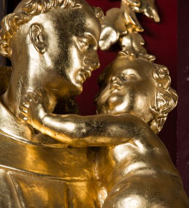 Zdjęcie nr 7: Rzeźba drążona, ustawiona na niskiej podstawie. Święty ukazany frontalnie, z głową skierowaną w lewo, w kontrapoście. Na lewym ramieniu podtrzymuje obejmujące go Dzieciątko Jezus, prawą opuszczoną wyciąga w bok. Twarz ma szczupłą, kwadratową, o podkreślonej linii żuchwy, migdałowatych, blisko siebie osadzonych oczach, małym nosie i rozchylonych ustach. Włosy ma krótkie, kręcone, na głowie tonsura. Święty ubrany jest w habit z kapturem, przepasany podwójnym szczurem ze zwisem z jednym guzem. Dzieciątko zwrócone w trzech czwartych w prawo, rączkami obejmuje głowę świętego, przepasane jest w partii podbrzusza pieluszką. Figury niemal w całości złocone, detale srebrzone.