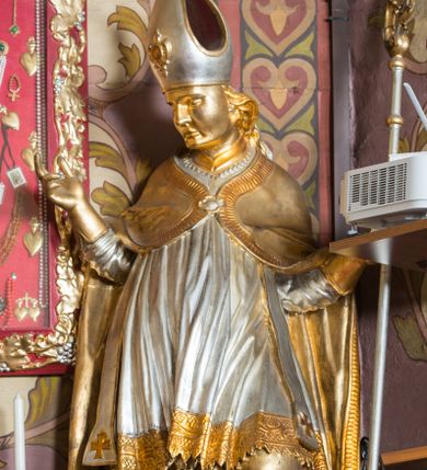 Zdjęcie nr 1: Rzeźba drążona, ustawiona na niskiej podstawie. Święty zwrócony w trzech czwartych w prawo, w kontrapoście, z pochyloną głową i lekko esowato wygiętą sylwetką. Obie ręce  ma rozłożone na boki, w lewej trzyma pastorał, prawą dłoń unosi w geście błogosławieństwa. Twarz ma szczupłą, kwadratową, o wyraźnie zarysowanej żuchwie, migdałowatych oczach, dużym nosie i wąskich, rozchylonych ustach; okoloną falowanymi włosami opadającymi na plecy. Na głowie ma infułę z taśmami (fannones). Święty ubrany jest w albę, rokietę dekorowaną płaskorzeźbioną koronką przy brzegu, stułę oraz kapę zapiętą klamrą na piersi, na dłoniach ma rękawiczki. Przy lewej nodze Stanisława popiersie mężczyzny ujęte tkaniną z głową skierowaną w górę. Figura niemal w całości złocona, detale stroju srebrzone.