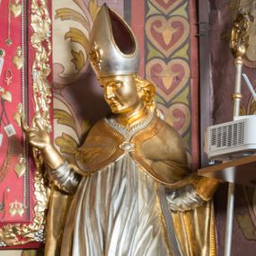 Zdjęcie nr 1: Rzeźba drążona, ustawiona na niskiej podstawie. Święty zwrócony w trzech czwartych w prawo, w kontrapoście, z pochyloną głową i lekko esowato wygiętą sylwetką. Obie ręce  ma rozłożone na boki, w lewej trzyma pastorał, prawą dłoń unosi w geście błogosławieństwa. Twarz ma szczupłą, kwadratową, o wyraźnie zarysowanej żuchwie, migdałowatych oczach, dużym nosie i wąskich, rozchylonych ustach; okoloną falowanymi włosami opadającymi na plecy. Na głowie ma infułę z taśmami (fannones). Święty ubrany jest w albę, rokietę dekorowaną płaskorzeźbioną koronką przy brzegu, stułę oraz kapę zapiętą klamrą na piersi, na dłoniach ma rękawiczki. Przy lewej nodze Stanisława popiersie mężczyzny ujęte tkaniną z głową skierowaną w górę. Figura niemal w całości złocona, detale stroju srebrzone.