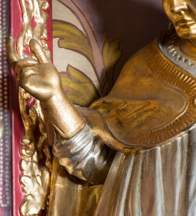 Zdjęcie nr 12: Rzeźba drążona, ustawiona na niskiej podstawie. Święty zwrócony w trzech czwartych w prawo, w kontrapoście, z pochyloną głową i lekko esowato wygiętą sylwetką. Obie ręce  ma rozłożone na boki, w lewej trzyma pastorał, prawą dłoń unosi w geście błogosławieństwa. Twarz ma szczupłą, kwadratową, o wyraźnie zarysowanej żuchwie, migdałowatych oczach, dużym nosie i wąskich, rozchylonych ustach; okoloną falowanymi włosami opadającymi na plecy. Na głowie ma infułę z taśmami (fannones). Święty ubrany jest w albę, rokietę dekorowaną płaskorzeźbioną koronką przy brzegu, stułę oraz kapę zapiętą klamrą na piersi, na dłoniach ma rękawiczki. Przy lewej nodze Stanisława popiersie mężczyzny ujęte tkaniną z głową skierowaną w górę. Figura niemal w całości złocona, detale stroju srebrzone.