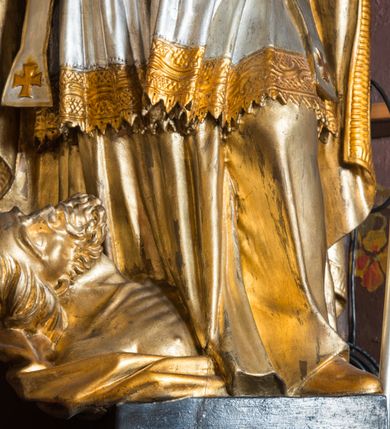 Zdjęcie nr 6: Rzeźba drążona, ustawiona na niskiej podstawie. Święty zwrócony w trzech czwartych w prawo, w kontrapoście, z pochyloną głową i lekko esowato wygiętą sylwetką. Obie ręce  ma rozłożone na boki, w lewej trzyma pastorał, prawą dłoń unosi w geście błogosławieństwa. Twarz ma szczupłą, kwadratową, o wyraźnie zarysowanej żuchwie, migdałowatych oczach, dużym nosie i wąskich, rozchylonych ustach; okoloną falowanymi włosami opadającymi na plecy. Na głowie ma infułę z taśmami (fannones). Święty ubrany jest w albę, rokietę dekorowaną płaskorzeźbioną koronką przy brzegu, stułę oraz kapę zapiętą klamrą na piersi, na dłoniach ma rękawiczki. Przy lewej nodze Stanisława popiersie mężczyzny ujęte tkaniną z głową skierowaną w górę. Figura niemal w całości złocona, detale stroju srebrzone.