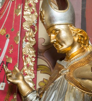 Zdjęcie nr 14: Rzeźba drążona, ustawiona na niskiej podstawie. Święty zwrócony w trzech czwartych w prawo, w kontrapoście, z pochyloną głową i lekko esowato wygiętą sylwetką. Obie ręce  ma rozłożone na boki, w lewej trzyma pastorał, prawą dłoń unosi w geście błogosławieństwa. Twarz ma szczupłą, kwadratową, o wyraźnie zarysowanej żuchwie, migdałowatych oczach, dużym nosie i wąskich, rozchylonych ustach; okoloną falowanymi włosami opadającymi na plecy. Na głowie ma infułę z taśmami (fannones). Święty ubrany jest w albę, rokietę dekorowaną płaskorzeźbioną koronką przy brzegu, stułę oraz kapę zapiętą klamrą na piersi, na dłoniach ma rękawiczki. Przy lewej nodze Stanisława popiersie mężczyzny ujęte tkaniną z głową skierowaną w górę. Figura niemal w całości złocona, detale stroju srebrzone.