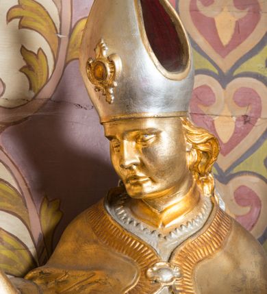 Zdjęcie nr 13: Rzeźba drążona, ustawiona na niskiej podstawie. Święty zwrócony w trzech czwartych w prawo, w kontrapoście, z pochyloną głową i lekko esowato wygiętą sylwetką. Obie ręce  ma rozłożone na boki, w lewej trzyma pastorał, prawą dłoń unosi w geście błogosławieństwa. Twarz ma szczupłą, kwadratową, o wyraźnie zarysowanej żuchwie, migdałowatych oczach, dużym nosie i wąskich, rozchylonych ustach; okoloną falowanymi włosami opadającymi na plecy. Na głowie ma infułę z taśmami (fannones). Święty ubrany jest w albę, rokietę dekorowaną płaskorzeźbioną koronką przy brzegu, stułę oraz kapę zapiętą klamrą na piersi, na dłoniach ma rękawiczki. Przy lewej nodze Stanisława popiersie mężczyzny ujęte tkaniną z głową skierowaną w górę. Figura niemal w całości złocona, detale stroju srebrzone.