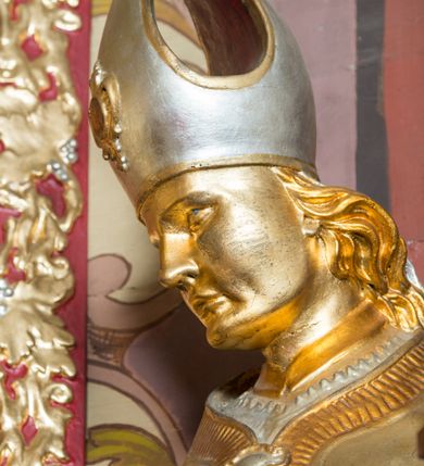 Zdjęcie nr 15: Rzeźba drążona, ustawiona na niskiej podstawie. Święty zwrócony w trzech czwartych w prawo, w kontrapoście, z pochyloną głową i lekko esowato wygiętą sylwetką. Obie ręce  ma rozłożone na boki, w lewej trzyma pastorał, prawą dłoń unosi w geście błogosławieństwa. Twarz ma szczupłą, kwadratową, o wyraźnie zarysowanej żuchwie, migdałowatych oczach, dużym nosie i wąskich, rozchylonych ustach; okoloną falowanymi włosami opadającymi na plecy. Na głowie ma infułę z taśmami (fannones). Święty ubrany jest w albę, rokietę dekorowaną płaskorzeźbioną koronką przy brzegu, stułę oraz kapę zapiętą klamrą na piersi, na dłoniach ma rękawiczki. Przy lewej nodze Stanisława popiersie mężczyzny ujęte tkaniną z głową skierowaną w górę. Figura niemal w całości złocona, detale stroju srebrzone.