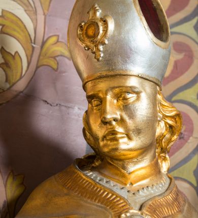 Zdjęcie nr 16: Rzeźba drążona, ustawiona na niskiej podstawie. Święty zwrócony w trzech czwartych w prawo, w kontrapoście, z pochyloną głową i lekko esowato wygiętą sylwetką. Obie ręce  ma rozłożone na boki, w lewej trzyma pastorał, prawą dłoń unosi w geście błogosławieństwa. Twarz ma szczupłą, kwadratową, o wyraźnie zarysowanej żuchwie, migdałowatych oczach, dużym nosie i wąskich, rozchylonych ustach; okoloną falowanymi włosami opadającymi na plecy. Na głowie ma infułę z taśmami (fannones). Święty ubrany jest w albę, rokietę dekorowaną płaskorzeźbioną koronką przy brzegu, stułę oraz kapę zapiętą klamrą na piersi, na dłoniach ma rękawiczki. Przy lewej nodze Stanisława popiersie mężczyzny ujęte tkaniną z głową skierowaną w górę. Figura niemal w całości złocona, detale stroju srebrzone.