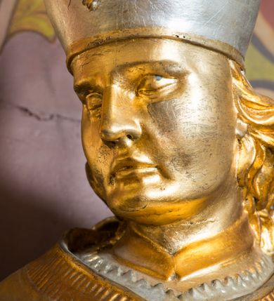 Zdjęcie nr 18: Rzeźba drążona, ustawiona na niskiej podstawie. Święty zwrócony w trzech czwartych w prawo, w kontrapoście, z pochyloną głową i lekko esowato wygiętą sylwetką. Obie ręce  ma rozłożone na boki, w lewej trzyma pastorał, prawą dłoń unosi w geście błogosławieństwa. Twarz ma szczupłą, kwadratową, o wyraźnie zarysowanej żuchwie, migdałowatych oczach, dużym nosie i wąskich, rozchylonych ustach; okoloną falowanymi włosami opadającymi na plecy. Na głowie ma infułę z taśmami (fannones). Święty ubrany jest w albę, rokietę dekorowaną płaskorzeźbioną koronką przy brzegu, stułę oraz kapę zapiętą klamrą na piersi, na dłoniach ma rękawiczki. Przy lewej nodze Stanisława popiersie mężczyzny ujęte tkaniną z głową skierowaną w górę. Figura niemal w całości złocona, detale stroju srebrzone.