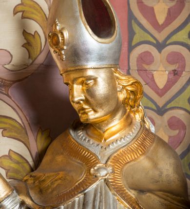 Zdjęcie nr 11: Rzeźba drążona, ustawiona na niskiej podstawie. Święty zwrócony w trzech czwartych w prawo, w kontrapoście, z pochyloną głową i lekko esowato wygiętą sylwetką. Obie ręce  ma rozłożone na boki, w lewej trzyma pastorał, prawą dłoń unosi w geście błogosławieństwa. Twarz ma szczupłą, kwadratową, o wyraźnie zarysowanej żuchwie, migdałowatych oczach, dużym nosie i wąskich, rozchylonych ustach; okoloną falowanymi włosami opadającymi na plecy. Na głowie ma infułę z taśmami (fannones). Święty ubrany jest w albę, rokietę dekorowaną płaskorzeźbioną koronką przy brzegu, stułę oraz kapę zapiętą klamrą na piersi, na dłoniach ma rękawiczki. Przy lewej nodze Stanisława popiersie mężczyzny ujęte tkaniną z głową skierowaną w górę. Figura niemal w całości złocona, detale stroju srebrzone.