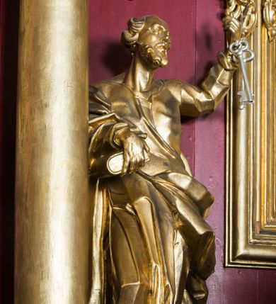 Zdjęcie nr 8: Rzeźba ścięta z tyłu i drążona, ustawiona na niskiej podstawie. Święty zwrócony frontalnie, z uniesioną głową skierowaną w lewo, w kontrapoście. Prawą ręką przytrzymuje na biodrze księgę, lewą unosi do góry w geście wskazującym dłoni, na której ma zawieszona dwa klucze. Twarz ma szczupłą, pociągłą, z wysokim czołem, oczy okrągłe, wyłupiaste, głęboko osadzone, nos garbaty, usta pełne i rozchylone. Twarz okolona krótką, gęstą brodą, włosy skręcone w pojedyncze pukle po bokach głowy, na jej czubku łysina. Święty ubrany jest w tunikę, ściśle przylegającą do ciała, marszczoną i łamaną, przepasaną w talii oraz płaszcz przerzucony przez prawe przedramię, wywinięty na lewym boku i podwieszony na prawym. Figura w całości złocona, klucze srebrzone.