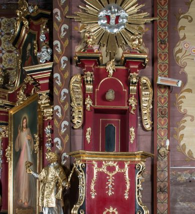 Zdjęcie nr 2: Ambona przyścienna, wisząca, z dostępem od strony zakrystii. Kosz na planie kwadratu o ściętych narożach, na podstawie w formie wałka z wyłamanymi, ściętymi narożami, dekorowanego rokokowymi kartuszami; z podwieszeniem w formie stożka o wklęsłych bokach ze spływami wolutowymi w narożach, zakończonym szyszką. Kosz o ściętych narożach ujęty pilastrami, których trzony przechodzą w spływy wolutowe dekorowane kwiatowymi zwisami, podtrzymującymi gzyms stanowiący parapet; ścianki dekorowane rocaille&#039;em z kwiatowymi zwisami. Zaplecek wąski, ujęty pilastrami dźwigającymi silnie wysunięte imposty podtrzymywane przez konsole, na nich wazony. Trzony dekorowane rokokowymi kampanulami oraz rocaille&#039;em. W polu wnęka z wąskimi drzwiami, dekorowanymi płycinami; ponad nimi płycina i rocaille. Zaplecek ujęty uszami utworzonymi z rocaille&#039;u. Całość zwieńczona glorią promienistą, w której centrum znajduje się gołębica Ducha Świętego w otoku z obłoków. Struktura polichromowana w kolorze czerwonym z czarnymi detalami, profile i ornamentyka złocone i srebrzone.