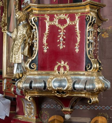 Zdjęcie nr 4: Ambona przyścienna, wisząca, z dostępem od strony zakrystii. Kosz na planie kwadratu o ściętych narożach, na podstawie w formie wałka z wyłamanymi, ściętymi narożami, dekorowanego rokokowymi kartuszami; z podwieszeniem w formie stożka o wklęsłych bokach ze spływami wolutowymi w narożach, zakończonym szyszką. Kosz o ściętych narożach ujęty pilastrami, których trzony przechodzą w spływy wolutowe dekorowane kwiatowymi zwisami, podtrzymującymi gzyms stanowiący parapet; ścianki dekorowane rocaille'em z kwiatowymi zwisami. Zaplecek wąski, ujęty pilastrami dźwigającymi silnie wysunięte imposty podtrzymywane przez konsole, na nich wazony. Trzony dekorowane rokokowymi kampanulami oraz rocaille'em. W polu wnęka z wąskimi drzwiami, dekorowanymi płycinami; ponad nimi płycina i rocaille. Zaplecek ujęty uszami utworzonymi z rocaille'u. Całość zwieńczona glorią promienistą, w której centrum znajduje się gołębica Ducha Świętego w otoku z obłoków. Struktura polichromowana w kolorze czerwonym z czarnymi detalami, profile i ornamentyka złocone i srebrzone.