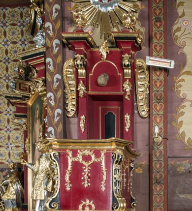Zdjęcie nr 1: Ambona przyścienna, wisząca, z dostępem od strony zakrystii. Kosz na planie kwadratu o ściętych narożach, na podstawie w formie wałka z wyłamanymi, ściętymi narożami, dekorowanego rokokowymi kartuszami; z podwieszeniem w formie stożka o wklęsłych bokach ze spływami wolutowymi w narożach, zakończonym szyszką. Kosz o ściętych narożach ujęty pilastrami, których trzony przechodzą w spływy wolutowe dekorowane kwiatowymi zwisami, podtrzymującymi gzyms stanowiący parapet; ścianki dekorowane rocaille'em z kwiatowymi zwisami. Zaplecek wąski, ujęty pilastrami dźwigającymi silnie wysunięte imposty podtrzymywane przez konsole, na nich wazony. Trzony dekorowane rokokowymi kampanulami oraz rocaille'em. W polu wnęka z wąskimi drzwiami, dekorowanymi płycinami; ponad nimi płycina i rocaille. Zaplecek ujęty uszami utworzonymi z rocaille'u. Całość zwieńczona glorią promienistą, w której centrum znajduje się gołębica Ducha Świętego w otoku z obłoków. Struktura polichromowana w kolorze czerwonym z czarnymi detalami, profile i ornamentyka złocone i srebrzone.