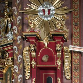 Zdjęcie nr 1: Ambona przyścienna, wisząca, z dostępem od strony zakrystii. Kosz na planie kwadratu o ściętych narożach, na podstawie w formie wałka z wyłamanymi, ściętymi narożami, dekorowanego rokokowymi kartuszami; z podwieszeniem w formie stożka o wklęsłych bokach ze spływami wolutowymi w narożach, zakończonym szyszką. Kosz o ściętych narożach ujęty pilastrami, których trzony przechodzą w spływy wolutowe dekorowane kwiatowymi zwisami, podtrzymującymi gzyms stanowiący parapet; ścianki dekorowane rocaille'em z kwiatowymi zwisami. Zaplecek wąski, ujęty pilastrami dźwigającymi silnie wysunięte imposty podtrzymywane przez konsole, na nich wazony. Trzony dekorowane rokokowymi kampanulami oraz rocaille'em. W polu wnęka z wąskimi drzwiami, dekorowanymi płycinami; ponad nimi płycina i rocaille. Zaplecek ujęty uszami utworzonymi z rocaille'u. Całość zwieńczona glorią promienistą, w której centrum znajduje się gołębica Ducha Świętego w otoku z obłoków. Struktura polichromowana w kolorze czerwonym z czarnymi detalami, profile i ornamentyka złocone i srebrzone.