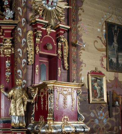Zdjęcie nr 3: Ambona przyścienna, wisząca, z dostępem od strony zakrystii. Kosz na planie kwadratu o ściętych narożach, na podstawie w formie wałka z wyłamanymi, ściętymi narożami, dekorowanego rokokowymi kartuszami; z podwieszeniem w formie stożka o wklęsłych bokach ze spływami wolutowymi w narożach, zakończonym szyszką. Kosz o ściętych narożach ujęty pilastrami, których trzony przechodzą w spływy wolutowe dekorowane kwiatowymi zwisami, podtrzymującymi gzyms stanowiący parapet; ścianki dekorowane rocaille&#039;em z kwiatowymi zwisami. Zaplecek wąski, ujęty pilastrami dźwigającymi silnie wysunięte imposty podtrzymywane przez konsole, na nich wazony. Trzony dekorowane rokokowymi kampanulami oraz rocaille&#039;em. W polu wnęka z wąskimi drzwiami, dekorowanymi płycinami; ponad nimi płycina i rocaille. Zaplecek ujęty uszami utworzonymi z rocaille&#039;u. Całość zwieńczona glorią promienistą, w której centrum znajduje się gołębica Ducha Świętego w otoku z obłoków. Struktura polichromowana w kolorze czerwonym z czarnymi detalami, profile i ornamentyka złocone i srebrzone.