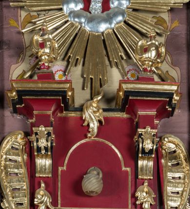 Zdjęcie nr 5: Ambona przyścienna, wisząca, z dostępem od strony zakrystii. Kosz na planie kwadratu o ściętych narożach, na podstawie w formie wałka z wyłamanymi, ściętymi narożami, dekorowanego rokokowymi kartuszami; z podwieszeniem w formie stożka o wklęsłych bokach ze spływami wolutowymi w narożach, zakończonym szyszką. Kosz o ściętych narożach ujęty pilastrami, których trzony przechodzą w spływy wolutowe dekorowane kwiatowymi zwisami, podtrzymującymi gzyms stanowiący parapet; ścianki dekorowane rocaille&#039;em z kwiatowymi zwisami. Zaplecek wąski, ujęty pilastrami dźwigającymi silnie wysunięte imposty podtrzymywane przez konsole, na nich wazony. Trzony dekorowane rokokowymi kampanulami oraz rocaille&#039;em. W polu wnęka z wąskimi drzwiami, dekorowanymi płycinami; ponad nimi płycina i rocaille. Zaplecek ujęty uszami utworzonymi z rocaille&#039;u. Całość zwieńczona glorią promienistą, w której centrum znajduje się gołębica Ducha Świętego w otoku z obłoków. Struktura polichromowana w kolorze czerwonym z czarnymi detalami, profile i ornamentyka złocone i srebrzone.