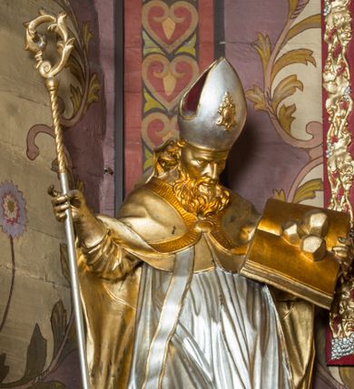Zdjęcie nr 1: Rzeźba ścięta z tyłu i drążona, ustawiona na niskiej podstawie. Święty zwrócony w trzech czwartych w lewo, z pochyloną głową, w kontrapoście i lekko esowato wygiętą sylwetką. W prawej, wysuniętej w bok ręce trzyma pastorał, lewą podtrzymuje otwartą księgę, na której leżą trzy kule. Twarz ma szczupłą, pociągłą, o podkreślonych kościach policzkowych, blisko osadzonych, migdałowatych oczach, dużym nosie i wąskich ustach. Twarz okolona jest gęstą brodą opadającą na pierś, rzeźbioną w drobne pukle oraz półdługimi, kręconymi włosami opadającymi w na tył głowy. Na głowie ma infułę z taśmami (fannones). Święty ubrany jest w albę, rokietę dekorowaną przy brzegu płaskorzeźbioną koronką, mucet, stułę, kapę zapiętą na piersi, na dłoniach ma rękawiczki. Przy lewej nodze szaflik z trzema półpostaciami w środku z głowami skierowanymi w górę i rękami złożonymi w geście modlitwy. Figura niemal w całości złocona, detale stroju srebrzone, szaflik polichromowany na czerwono. 