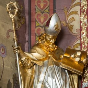 Zdjęcie nr 1: Rzeźba ścięta z tyłu i drążona, ustawiona na niskiej podstawie. Święty zwrócony w trzech czwartych w lewo, z pochyloną głową, w kontrapoście i lekko esowato wygiętą sylwetką. W prawej, wysuniętej w bok ręce trzyma pastorał, lewą podtrzymuje otwartą księgę, na której leżą trzy kule. Twarz ma szczupłą, pociągłą, o podkreślonych kościach policzkowych, blisko osadzonych, migdałowatych oczach, dużym nosie i wąskich ustach. Twarz okolona jest gęstą brodą opadającą na pierś, rzeźbioną w drobne pukle oraz półdługimi, kręconymi włosami opadającymi w na tył głowy. Na głowie ma infułę z taśmami (fannones). Święty ubrany jest w albę, rokietę dekorowaną przy brzegu płaskorzeźbioną koronką, mucet, stułę, kapę zapiętą na piersi, na dłoniach ma rękawiczki. Przy lewej nodze szaflik z trzema półpostaciami w środku z głowami skierowanymi w górę i rękami złożonymi w geście modlitwy. Figura niemal w całości złocona, detale stroju srebrzone, szaflik polichromowany na czerwono. 