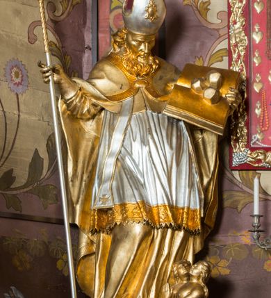 Zdjęcie nr 8: Rzeźba ścięta z tyłu i drążona, ustawiona na niskiej podstawie. Święty zwrócony w trzech czwartych w lewo, z pochyloną głową, w kontrapoście i lekko esowato wygiętą sylwetką. W prawej, wysuniętej w bok ręce trzyma pastorał, lewą podtrzymuje otwartą księgę, na której leżą trzy kule. Twarz ma szczupłą, pociągłą, o podkreślonych kościach policzkowych, blisko osadzonych, migdałowatych oczach, dużym nosie i wąskich ustach. Twarz okolona jest gęstą brodą opadającą na pierś, rzeźbioną w drobne pukle oraz półdługimi, kręconymi włosami opadającymi w na tył głowy. Na głowie ma infułę z taśmami (fannones). Święty ubrany jest w albę, rokietę dekorowaną przy brzegu płaskorzeźbioną koronką, mucet, stułę, kapę zapiętą na piersi, na dłoniach ma rękawiczki. Przy lewej nodze szaflik z trzema półpostaciami w środku z głowami skierowanymi w górę i rękami złożonymi w geście modlitwy. Figura niemal w całości złocona, detale stroju srebrzone, szaflik polichromowany na czerwono. 