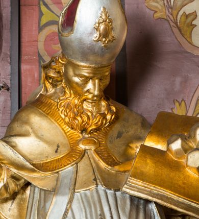 Zdjęcie nr 9: Rzeźba ścięta z tyłu i drążona, ustawiona na niskiej podstawie. Święty zwrócony w trzech czwartych w lewo, z pochyloną głową, w kontrapoście i lekko esowato wygiętą sylwetką. W prawej, wysuniętej w bok ręce trzyma pastorał, lewą podtrzymuje otwartą księgę, na której leżą trzy kule. Twarz ma szczupłą, pociągłą, o podkreślonych kościach policzkowych, blisko osadzonych, migdałowatych oczach, dużym nosie i wąskich ustach. Twarz okolona jest gęstą brodą opadającą na pierś, rzeźbioną w drobne pukle oraz półdługimi, kręconymi włosami opadającymi w na tył głowy. Na głowie ma infułę z taśmami (fannones). Święty ubrany jest w albę, rokietę dekorowaną przy brzegu płaskorzeźbioną koronką, mucet, stułę, kapę zapiętą na piersi, na dłoniach ma rękawiczki. Przy lewej nodze szaflik z trzema półpostaciami w środku z głowami skierowanymi w górę i rękami złożonymi w geście modlitwy. Figura niemal w całości złocona, detale stroju srebrzone, szaflik polichromowany na czerwono. 