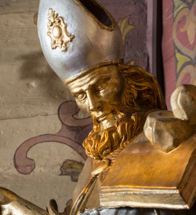 Zdjęcie nr 15: Rzeźba ścięta z tyłu i drążona, ustawiona na niskiej podstawie. Święty zwrócony w trzech czwartych w lewo, z pochyloną głową, w kontrapoście i lekko esowato wygiętą sylwetką. W prawej, wysuniętej w bok ręce trzyma pastorał, lewą podtrzymuje otwartą księgę, na której leżą trzy kule. Twarz ma szczupłą, pociągłą, o podkreślonych kościach policzkowych, blisko osadzonych, migdałowatych oczach, dużym nosie i wąskich ustach. Twarz okolona jest gęstą brodą opadającą na pierś, rzeźbioną w drobne pukle oraz półdługimi, kręconymi włosami opadającymi w na tył głowy. Na głowie ma infułę z taśmami (fannones). Święty ubrany jest w albę, rokietę dekorowaną przy brzegu płaskorzeźbioną koronką, mucet, stułę, kapę zapiętą na piersi, na dłoniach ma rękawiczki. Przy lewej nodze szaflik z trzema półpostaciami w środku z głowami skierowanymi w górę i rękami złożonymi w geście modlitwy. Figura niemal w całości złocona, detale stroju srebrzone, szaflik polichromowany na czerwono. 