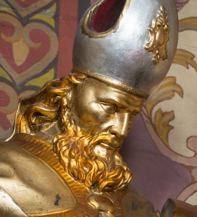 Zdjęcie nr 3: Rzeźba ścięta z tyłu i drążona, ustawiona na niskiej podstawie. Święty zwrócony w trzech czwartych w lewo, z pochyloną głową, w kontrapoście i lekko esowato wygiętą sylwetką. W prawej, wysuniętej w bok ręce trzyma pastorał, lewą podtrzymuje otwartą księgę, na której leżą trzy kule. Twarz ma szczupłą, pociągłą, o podkreślonych kościach policzkowych, blisko osadzonych, migdałowatych oczach, dużym nosie i wąskich ustach. Twarz okolona jest gęstą brodą opadającą na pierś, rzeźbioną w drobne pukle oraz półdługimi, kręconymi włosami opadającymi w na tył głowy. Na głowie ma infułę z taśmami (fannones). Święty ubrany jest w albę, rokietę dekorowaną przy brzegu płaskorzeźbioną koronką, mucet, stułę, kapę zapiętą na piersi, na dłoniach ma rękawiczki. Przy lewej nodze szaflik z trzema półpostaciami w środku z głowami skierowanymi w górę i rękami złożonymi w geście modlitwy. Figura niemal w całości złocona, detale stroju srebrzone, szaflik polichromowany na czerwono. 