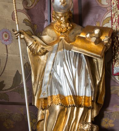 Zdjęcie nr 5: Rzeźba ścięta z tyłu i drążona, ustawiona na niskiej podstawie. Święty zwrócony w trzech czwartych w lewo, z pochyloną głową, w kontrapoście i lekko esowato wygiętą sylwetką. W prawej, wysuniętej w bok ręce trzyma pastorał, lewą podtrzymuje otwartą księgę, na której leżą trzy kule. Twarz ma szczupłą, pociągłą, o podkreślonych kościach policzkowych, blisko osadzonych, migdałowatych oczach, dużym nosie i wąskich ustach. Twarz okolona jest gęstą brodą opadającą na pierś, rzeźbioną w drobne pukle oraz półdługimi, kręconymi włosami opadającymi w na tył głowy. Na głowie ma infułę z taśmami (fannones). Święty ubrany jest w albę, rokietę dekorowaną przy brzegu płaskorzeźbioną koronką, mucet, stułę, kapę zapiętą na piersi, na dłoniach ma rękawiczki. Przy lewej nodze szaflik z trzema półpostaciami w środku z głowami skierowanymi w górę i rękami złożonymi w geście modlitwy. Figura niemal w całości złocona, detale stroju srebrzone, szaflik polichromowany na czerwono. 