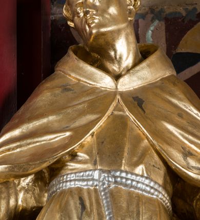 Zdjęcie nr 11: Rzeźba drążona, ustawiona na niewielkiej podstawie. Święty ukazany frontalnie z uniesioną głową, skierowaną w prawo, w kontrapoście, odchylony w tył. Obie ręce ma rozłożone na boki, a dłonie skierowane w prawo. Twarz ma szczupłą, kwadratową, z wyraźnie zarysowaną żuchwą, o migdałowatych, blisko siebie osadzonych oczach, małym nosie i ustach. Na głowie ma tonsurę, wokół krótkie, kręcone włosy. Ubrany jest w habit, ściśle przylegający do ciała, przepasany w talii podwójnym szczurem z końcem z jednym guzem oraz krótki płaszcz z kapturem zapięty na piersi i opadający na ramiona. Figura niemal w całości złocona, sznur srebrzony.