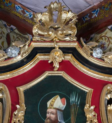 Zdjęcie nr 25: Nastawa architektoniczna, jednoosiowa, jednokondygnacyjna ze zwieńczeniem, na wysokim cokole z wyodrębnionymi konsolami. Ołtarz drewniany, sarkofagowy, na cokole, zwężający się ku dołowi z drewnianą mensą i kamiennym portatylem. Nastawa ujęta parą zdwojonych pilastrów zwróconych pod kątem na zewnątrz, dekorowanych zwisami kwiatowymi, dźwigających silnie wysunięte imposty podtrzymywane przez konsole. Na tle pilastrów figury św. Wawrzyńca i św. Szczepana ustawione na konsolach. Całe pole środkowe wypełnia wtórnie umieszczony obraz „Jezu ufam Tobie” w profilowanej ramie zdobionej rocaille&#039;em. Nad nim fragmenty rocaille&#039;u, pod nim płycina z dekoracją roślinną. Zwieńczenie w formie trapezu ujętego spływami wolutowymi, dekorowanymi liśćmi palmowymi, ze ślimacznicami w kształcie meandru, spływającymi na belkowanie, na których stoją wazony. Całość zamknięta fragmentem wyłamanego trójbocznie gzymsu, zwieńczonego rokokowym szczytem z kwiatowymi gałązkami. W polu obraz św. Wojciecha w formie zbliżonej do zwieńczenia, ujęty ramą dekorowaną rocaille&#039;em. Struktura polichromowana w kolorze głębokiej czerwieni, z czarnymi profilami oraz złoconymi ramami, profilami i ornamentyką.
