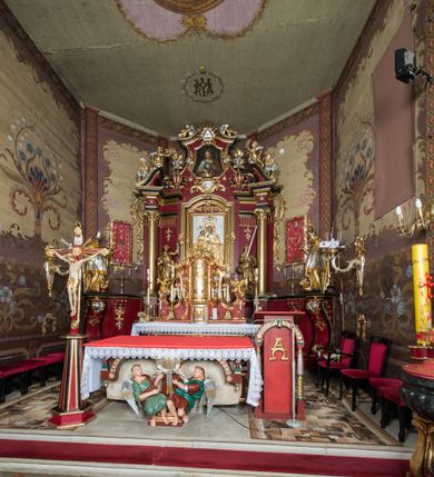Zdjęcie nr 35: Nastawa architektoniczna, jednoosiowa, jednokondygnacyjna ze zwieńczeniem, ustawiona na dwustrefowym cokole z wyodrębnionymi prostopadłościanami. Ołtarz drewniany, prostopadłościenny z drewnianą mensą. Antepedium wydzielone ramą i wysunięte do przodu, ujęte pilastrami dźwigającymi niepełne belkowanie, w polu wieniec laurowy z rozetą, nad nim kokarda z karbowanej wstęgi flankowana przez dwa podwieszone festony z tkaniny, poniżej wić roślinna. Na ołtarzu ustawione tabernakulum w formie metalowej skrzynki w drewnianej obudowie, ujęte ustawionymi pod kątem spływami wolutowymi. Ścianka frontowa wypukła, z drewnianymi drzwiczkami w kształcie stojącego prostokąta zamkniętego łukiem półkolistym z płaskorzeźbionym kielichem z hostią i kłosami zbóż. Na nim wysoki tron wystawienia z wypukłym frontem z zamontowanym wtórnie nowym tabernakulum (wcześniej obrotowa nisza). Tron ujęty pilastrami ustawionymi pod kątem, o trzonach przechodzących w spływy wolutowe, dekorowane zwisami kwiatowymi; zwieńczony jest trzema rocaille&#039;owymi kabłąkami dekorowanymi zwisami kwiatowymi, podtrzymującymi figurkę pelikana karmiącego młode w gnieździe. Tabernakulum i tron flankowane spływami z podwieszonymi zwisami kwiatowymi zakończonymi cokołami, dźwigającymi przerwany wolutowy przyczółek z figurkami adorujących aniołów; spływy w górnej części zamknięte fragmentami gzymsu z palmetami. 
Nastawa ujęta dwiema parami pilastrów oraz stojącą na ich tle parą kolumn na cokołach dźwigających pełne belkowanie przerwane nad polem środkowym oraz fragmenty przerwanego, wolutowego przyczółka. Przed wewnętrzną parą pilastrów ustawione figury św. Piotra i św. Pawła; na belkowaniu wazony, na przyczółku putta trzymające słońce i księżyc. W polu środkowym podwójna wnęka w formie stojącego prostokąta zamkniętego łukiem odcinkowym nadwieszonym, ujęta profilowaną ramą, ze znacznie mniejszym obrazem Matki Boskiej w kształcie stojącego prostokąta. Nad nią medalion utworzony z wolut i zwisów, zwieńczony koroną otwartą. Zwieńczenie nastawy w formie trapezu ujętego wolutami spływającymi na belkowanie, zamknięte fragmentem falistego gzymsu, dekorowanym medalionem z Okiem Opatrzności w otoku z obłoków w glorii promienistej oraz gałązkami kwiatowymi. W polu obraz św. Katarzyny w formie zbliżonej do zwieńczenia, o ściętych dolnych narożach, w profilowanej ramie, z gałązkami kwiatowymi. 
Nastawa flankowana uszami utworzonymi z rocaille&#039;u, przytwierdzonymi do ściany. Ujęta parą wsporników na trumienki relikwiarzowe oraz parą dużych konsol dekorowanych rocaille&#039;m z figurami św. Mikołaja i św. Stanisława. Struktura polichromowana w kolorze głębokiej czerwieni z czarnymi profilami; kolumny, ornamentyka i profile złocone i srebrzone.