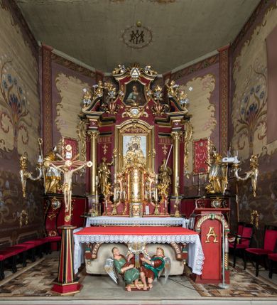 Zdjęcie nr 13: Nastawa architektoniczna, jednoosiowa, jednokondygnacyjna ze zwieńczeniem, ustawiona na dwustrefowym cokole z wyodrębnionymi prostopadłościanami. Ołtarz drewniany, prostopadłościenny z drewnianą mensą. Antepedium wydzielone ramą i wysunięte do przodu, ujęte pilastrami dźwigającymi niepełne belkowanie, w polu wieniec laurowy z rozetą, nad nim kokarda z karbowanej wstęgi flankowana przez dwa podwieszone festony z tkaniny, poniżej wić roślinna. Na ołtarzu ustawione tabernakulum w formie metalowej skrzynki w drewnianej obudowie, ujęte ustawionymi pod kątem spływami wolutowymi. Ścianka frontowa wypukła, z drewnianymi drzwiczkami w kształcie stojącego prostokąta zamkniętego łukiem półkolistym z płaskorzeźbionym kielichem z hostią i kłosami zbóż. Na nim wysoki tron wystawienia z wypukłym frontem z zamontowanym wtórnie nowym tabernakulum (wcześniej obrotowa nisza). Tron ujęty pilastrami ustawionymi pod kątem, o trzonach przechodzących w spływy wolutowe, dekorowane zwisami kwiatowymi; zwieńczony jest trzema rocaille&#039;owymi kabłąkami dekorowanymi zwisami kwiatowymi, podtrzymującymi figurkę pelikana karmiącego młode w gnieździe. Tabernakulum i tron flankowane spływami z podwieszonymi zwisami kwiatowymi zakończonymi cokołami, dźwigającymi przerwany wolutowy przyczółek z figurkami adorujących aniołów; spływy w górnej części zamknięte fragmentami gzymsu z palmetami. 
Nastawa ujęta dwiema parami pilastrów oraz stojącą na ich tle parą kolumn na cokołach dźwigających pełne belkowanie przerwane nad polem środkowym oraz fragmenty przerwanego, wolutowego przyczółka. Przed wewnętrzną parą pilastrów ustawione figury św. Piotra i św. Pawła; na belkowaniu wazony, na przyczółku putta trzymające słońce i księżyc. W polu środkowym podwójna wnęka w formie stojącego prostokąta zamkniętego łukiem odcinkowym nadwieszonym, ujęta profilowaną ramą, ze znacznie mniejszym obrazem Matki Boskiej w kształcie stojącego prostokąta. Nad nią medalion utworzony z wolut i zwisów, zwieńczony koroną otwartą. Zwieńczenie nastawy w formie trapezu ujętego wolutami spływającymi na belkowanie, zamknięte fragmentem falistego gzymsu, dekorowanym medalionem z Okiem Opatrzności w otoku z obłoków w glorii promienistej oraz gałązkami kwiatowymi. W polu obraz św. Katarzyny w formie zbliżonej do zwieńczenia, o ściętych dolnych narożach, w profilowanej ramie, z gałązkami kwiatowymi. 
Nastawa flankowana uszami utworzonymi z rocaille&#039;u, przytwierdzonymi do ściany. Ujęta parą wsporników na trumienki relikwiarzowe oraz parą dużych konsol dekorowanych rocaille&#039;m z figurami św. Mikołaja i św. Stanisława. Struktura polichromowana w kolorze głębokiej czerwieni z czarnymi profilami; kolumny, ornamentyka i profile złocone i srebrzone.