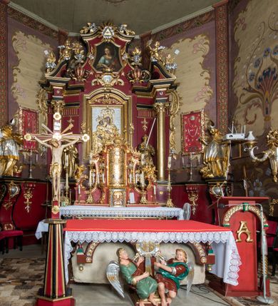 Zdjęcie nr 24: Nastawa architektoniczna, jednoosiowa, jednokondygnacyjna ze zwieńczeniem, ustawiona na dwustrefowym cokole z wyodrębnionymi prostopadłościanami. Ołtarz drewniany, prostopadłościenny z drewnianą mensą. Antepedium wydzielone ramą i wysunięte do przodu, ujęte pilastrami dźwigającymi niepełne belkowanie, w polu wieniec laurowy z rozetą, nad nim kokarda z karbowanej wstęgi flankowana przez dwa podwieszone festony z tkaniny, poniżej wić roślinna. Na ołtarzu ustawione tabernakulum w formie metalowej skrzynki w drewnianej obudowie, ujęte ustawionymi pod kątem spływami wolutowymi. Ścianka frontowa wypukła, z drewnianymi drzwiczkami w kształcie stojącego prostokąta zamkniętego łukiem półkolistym z płaskorzeźbionym kielichem z hostią i kłosami zbóż. Na nim wysoki tron wystawienia z wypukłym frontem z zamontowanym wtórnie nowym tabernakulum (wcześniej obrotowa nisza). Tron ujęty pilastrami ustawionymi pod kątem, o trzonach przechodzących w spływy wolutowe, dekorowane zwisami kwiatowymi; zwieńczony jest trzema rocaille'owymi kabłąkami dekorowanymi zwisami kwiatowymi, podtrzymującymi figurkę pelikana karmiącego młode w gnieździe. Tabernakulum i tron flankowane spływami z podwieszonymi zwisami kwiatowymi zakończonymi cokołami, dźwigającymi przerwany wolutowy przyczółek z figurkami adorujących aniołów; spływy w górnej części zamknięte fragmentami gzymsu z palmetami. 
Nastawa ujęta dwiema parami pilastrów oraz stojącą na ich tle parą kolumn na cokołach dźwigających pełne belkowanie przerwane nad polem środkowym oraz fragmenty przerwanego, wolutowego przyczółka. Przed wewnętrzną parą pilastrów ustawione figury św. Piotra i św. Pawła; na belkowaniu wazony, na przyczółku putta trzymające słońce i księżyc. W polu środkowym podwójna wnęka w formie stojącego prostokąta zamkniętego łukiem odcinkowym nadwieszonym, ujęta profilowaną ramą, ze znacznie mniejszym obrazem Matki Boskiej w kształcie stojącego prostokąta. Nad nią medalion utworzony z wolut i zwisów, zwieńczony koroną otwartą. Zwieńczenie nastawy w formie trapezu ujętego wolutami spływającymi na belkowanie, zamknięte fragmentem falistego gzymsu, dekorowanym medalionem z Okiem Opatrzności w otoku z obłoków w glorii promienistej oraz gałązkami kwiatowymi. W polu obraz św. Katarzyny w formie zbliżonej do zwieńczenia, o ściętych dolnych narożach, w profilowanej ramie, z gałązkami kwiatowymi. 
Nastawa flankowana uszami utworzonymi z rocaille'u, przytwierdzonymi do ściany. Ujęta parą wsporników na trumienki relikwiarzowe oraz parą dużych konsol dekorowanych rocaille'm z figurami św. Mikołaja i św. Stanisława. Struktura polichromowana w kolorze głębokiej czerwieni z czarnymi profilami; kolumny, ornamentyka i profile złocone i srebrzone.