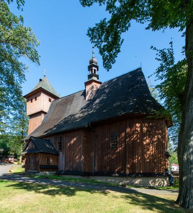 Zdjęcie nr 30: Kościół parafialny w Gruszowie znajduje się na niewielkim wzniesieniu w centrum miejscowości. Budowla jest orientowana, jednonawowa, z szerokim zamkniętym trójbocznie prezbiterium oraz czworoboczną wieżą od zachodu. Korpusu nawowy i prezbiterium charakteryzuje konstrukcja zrębowa, natomiast wieżę słupowa. Od północy do prezbiterium przylega zakrystia na rzucie prostokąta, natomiast do nawy zamknięta trójbocznie kaplica boczna. Do wnętrza kościoła prowadzą dwie kruchty, jedna od strony południowej, druga przez wieżę od zachodniej. Nad korpusem nawowym i prezbiterium znajduje się jednokalenicowy dach kryty gontem, a na styku nawy i prezbiterium usytuowana jest drewniana wieżyczka na sygnaturkę nakryta baniastym hełmem z latarnią. Zakrystia przekryta jest dachem pulpitowym, natomiast kaplica boczna oraz kruchty dachami dwuspadowymi krytymi gontem. Izbicową, o pochyłych ścianach wieżę wieńczy okuty blachą dach namiotowy. Elewacje osłania szalunek z pionowych desek z tzw. listwowaniem oraz pseudopilastry, dodatkowo wieżę w jednej trzeciej wysokości obiega niewielki parapet kryty gontem. W strukturze korpusu nawowego otwory okienne znajdują się po stronie południowej i mają kształt stojących prostokątów. W prezbiterium znajduje się po jednym oknie od południa i od wschodu, ich prostokątna forma jest zamknięta łukiem w ośli grzbiet. W kaplicy bocznej jest jedno okno od strony zachodniej. Ściana wschodnia zakrystii przepruta jest dwoma niewielkimi oknami, natomiast kruchty południowej jednym. W kruchcie zachodniej od południa jest jedno okno, natomiast w dolnej partii wieży znajdują się trzy, z czego dwa dolne oddziela od górnego niewielki daszek pulpitowy. Wnętrze korpusu nawowego i prezbiterium kościoła nakryte jest drewnianym stropem. Do kaplicy bocznej prowadzi szerokie przejście o ostrołukowym wykroju. Południowy portal w korpusie nawowym wieńczy wykrój w formie łuku w ośli grzbiet. Ściany i stropy wnętrza kościoła pokrywa secesyjna polichromia. 
Plac przykościelny otoczony jest drewnianym ogrodzeniem oraz starodrzewem.
