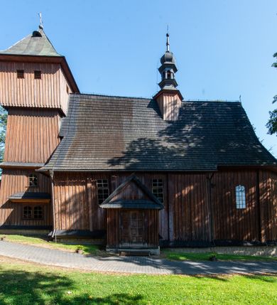 Zdjęcie nr 13: Kościół parafialny w Gruszowie znajduje się na niewielkim wzniesieniu w centrum miejscowości. Budowla jest orientowana, jednonawowa, z szerokim zamkniętym trójbocznie prezbiterium oraz czworoboczną wieżą od zachodu. Korpusu nawowy i prezbiterium charakteryzuje konstrukcja zrębowa, natomiast wieżę słupowa. Od północy do prezbiterium przylega zakrystia na rzucie prostokąta, natomiast do nawy zamknięta trójbocznie kaplica boczna. Do wnętrza kościoła prowadzą dwie kruchty, jedna od strony południowej, druga przez wieżę od zachodniej. Nad korpusem nawowym i prezbiterium znajduje się jednokalenicowy dach kryty gontem, a na styku nawy i prezbiterium usytuowana jest drewniana wieżyczka na sygnaturkę nakryta baniastym hełmem z latarnią. Zakrystia przekryta jest dachem pulpitowym, natomiast kaplica boczna oraz kruchty dachami dwuspadowymi krytymi gontem. Izbicową, o pochyłych ścianach wieżę wieńczy okuty blachą dach namiotowy. Elewacje osłania szalunek z pionowych desek z tzw. listwowaniem oraz pseudopilastry, dodatkowo wieżę w jednej trzeciej wysokości obiega niewielki parapet kryty gontem. W strukturze korpusu nawowego otwory okienne znajdują się po stronie południowej i mają kształt stojących prostokątów. W prezbiterium znajduje się po jednym oknie od południa i od wschodu, ich prostokątna forma jest zamknięta łukiem w ośli grzbiet. W kaplicy bocznej jest jedno okno od strony zachodniej. Ściana wschodnia zakrystii przepruta jest dwoma niewielkimi oknami, natomiast kruchty południowej jednym. W kruchcie zachodniej od południa jest jedno okno, natomiast w dolnej partii wieży znajdują się trzy, z czego dwa dolne oddziela od górnego niewielki daszek pulpitowy. Wnętrze korpusu nawowego i prezbiterium kościoła nakryte jest drewnianym stropem. Do kaplicy bocznej prowadzi szerokie przejście o ostrołukowym wykroju. Południowy portal w korpusie nawowym wieńczy wykrój w formie łuku w ośli grzbiet. Ściany i stropy wnętrza kościoła pokrywa secesyjna polichromia. 
Plac przykościelny otoczony jest drewnianym ogrodzeniem oraz starodrzewem.
