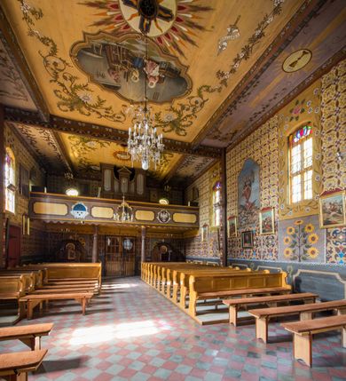 Zdjęcie nr 24: Kościół drewniany o konstrukcji zrębowej, elewacje jednolite, szalowane pionowo z listwowaniem, kruchta zachodnia o konstrukcji słupowo-ramowej. Więźba dachowa drewniana, krokwiowa. Budynek na kamiennym podmurowaniu przesłoniętym daszkiem gontowym. Kościół orientowany, jednonawowy, nawa na rzucie prostokąta, prezbiterium węższe, zakończone trójbocznie. Do wschodniej ściany prezbiterium przylega Ogrójec na rzucie prostokąta, od północy piętrowa zakrystia na planie prostokąta. Do południowej ściany nawy przylega niewielka kruchta na rzucie prostokąta oraz ołtarz polowy, od zachodu kruchta na planie prostokąta z węższym pięterkiem dzwonnym. Wnętrze podzielone lizenami, nakryte pułapami deskowymi. Pomiędzy nawą a prezbiterium prostokątny otwór tęczowy o zaokrąglonych narożach. Parapet chóru muzycznego w zachodniej części nawy podparty przez dwie kolumny o stylizowanych kapitelach. Otwory drzwiowe o prostokątnym wykroju w północnej ścianie prezbiterium, południowej i zachodniej nawy. Posadzka z płyt ceramicznych. Elewacja frontowa jednoosiowa. Na osi wejście do kruchty z dwuskrzydłowymi drzwiami, nakryte osobnym daszkiem dwuspadowym. Na północnej i południowej ścianie kruchty i pięterka dzwonnego niewielkie okna. Kruchta nakryta daszkiem trójspadowym gontowym, dzwonnica dwuspadowym, z krzyżem w partii szczytu. W elewacji południowej cztery otwory okienne w formie stojącego prostokąta zamkniętego łukiem półkolistym i odcinkowym, dwa w nawie i dwa w prezbiterium, w elewacji północnej dwa analogiczne otwory okienne. Na ścianie północnej zachowany fragment dawnych sobót, zabudowany pionowymi deskami. Na ścianie wschodniej prezbiterium Ogrójec w formie kapliczki przesłoniętej daszkiem dwuspadowym. Nawa nakryta dachem gontowym, dwuspadowym, półszczytowym, prezbiterium osobnym dachem wielospadowym, zakrystia przesłonięta dachem przyczółkowym, kruchta dwuspadowym, a ołtarz polowy trójspadowym. Nad nawą od strony prezbiterium wznosi się sześcioboczna wieżyczka na sygnaturkę z latarnią i hełmem cebulastym z kulą i krzyżem pokryta blachą. 



 
 
