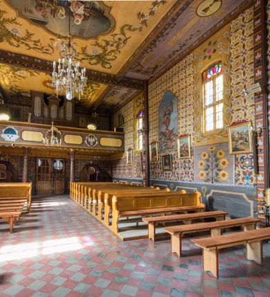 Zdjęcie nr 26: Kościół drewniany o konstrukcji zrębowej, elewacje jednolite, szalowane pionowo z listwowaniem, kruchta zachodnia o konstrukcji słupowo-ramowej. Więźba dachowa drewniana, krokwiowa. Budynek na kamiennym podmurowaniu przesłoniętym daszkiem gontowym. Kościół orientowany, jednonawowy, nawa na rzucie prostokąta, prezbiterium węższe, zakończone trójbocznie. Do wschodniej ściany prezbiterium przylega Ogrójec na rzucie prostokąta, od północy piętrowa zakrystia na planie prostokąta. Do południowej ściany nawy przylega niewielka kruchta na rzucie prostokąta oraz ołtarz polowy, od zachodu kruchta na planie prostokąta z węższym pięterkiem dzwonnym. Wnętrze podzielone lizenami, nakryte pułapami deskowymi. Pomiędzy nawą a prezbiterium prostokątny otwór tęczowy o zaokrąglonych narożach. Parapet chóru muzycznego w zachodniej części nawy podparty przez dwie kolumny o stylizowanych kapitelach. Otwory drzwiowe o prostokątnym wykroju w północnej ścianie prezbiterium, południowej i zachodniej nawy. Posadzka z płyt ceramicznych. Elewacja frontowa jednoosiowa. Na osi wejście do kruchty z dwuskrzydłowymi drzwiami, nakryte osobnym daszkiem dwuspadowym. Na północnej i południowej ścianie kruchty i pięterka dzwonnego niewielkie okna. Kruchta nakryta daszkiem trójspadowym gontowym, dzwonnica dwuspadowym, z krzyżem w partii szczytu. W elewacji południowej cztery otwory okienne w formie stojącego prostokąta zamkniętego łukiem półkolistym i odcinkowym, dwa w nawie i dwa w prezbiterium, w elewacji północnej dwa analogiczne otwory okienne. Na ścianie północnej zachowany fragment dawnych sobót, zabudowany pionowymi deskami. Na ścianie wschodniej prezbiterium Ogrójec w formie kapliczki przesłoniętej daszkiem dwuspadowym. Nawa nakryta dachem gontowym, dwuspadowym, półszczytowym, prezbiterium osobnym dachem wielospadowym, zakrystia przesłonięta dachem przyczółkowym, kruchta dwuspadowym, a ołtarz polowy trójspadowym. Nad nawą od strony prezbiterium wznosi się sześcioboczna wieżyczka na sygnaturkę z latarnią i hełmem cebulastym z kulą i krzyżem pokryta blachą. 



 
 
