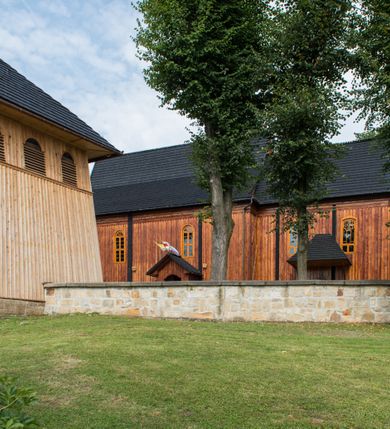 Zdjęcie nr 18: Kościół drewniany o konstrukcji zrębowej, elewacje jednolite, szalowane pionowo z listwowaniem, kruchta zachodnia o konstrukcji słupowo-ramowej. Więźba dachowa drewniana, krokwiowa. Budynek na kamiennym podmurowaniu przesłoniętym daszkiem gontowym. Kościół orientowany, jednonawowy, nawa na rzucie prostokąta, prezbiterium węższe, zakończone trójbocznie. Do wschodniej ściany prezbiterium przylega Ogrójec na rzucie prostokąta, od północy piętrowa zakrystia na planie prostokąta. Do południowej ściany nawy przylega niewielka kruchta na rzucie prostokąta oraz ołtarz polowy, od zachodu kruchta na planie prostokąta z węższym pięterkiem dzwonnym. Wnętrze podzielone lizenami, nakryte pułapami deskowymi. Pomiędzy nawą a prezbiterium prostokątny otwór tęczowy o zaokrąglonych narożach. Parapet chóru muzycznego w zachodniej części nawy podparty przez dwie kolumny o stylizowanych kapitelach. Otwory drzwiowe o prostokątnym wykroju w północnej ścianie prezbiterium, południowej i zachodniej nawy. Posadzka z płyt ceramicznych. Elewacja frontowa jednoosiowa. Na osi wejście do kruchty z dwuskrzydłowymi drzwiami, nakryte osobnym daszkiem dwuspadowym. Na północnej i południowej ścianie kruchty i pięterka dzwonnego niewielkie okna. Kruchta nakryta daszkiem trójspadowym gontowym, dzwonnica dwuspadowym, z krzyżem w partii szczytu. W elewacji południowej cztery otwory okienne w formie stojącego prostokąta zamkniętego łukiem półkolistym i odcinkowym, dwa w nawie i dwa w prezbiterium, w elewacji północnej dwa analogiczne otwory okienne. Na ścianie północnej zachowany fragment dawnych sobót, zabudowany pionowymi deskami. Na ścianie wschodniej prezbiterium Ogrójec w formie kapliczki przesłoniętej daszkiem dwuspadowym. Nawa nakryta dachem gontowym, dwuspadowym, półszczytowym, prezbiterium osobnym dachem wielospadowym, zakrystia przesłonięta dachem przyczółkowym, kruchta dwuspadowym, a ołtarz polowy trójspadowym. Nad nawą od strony prezbiterium wznosi się sześcioboczna wieżyczka na sygnaturkę z latarnią i hełmem cebulastym z kulą i krzyżem pokryta blachą. 



 
 
